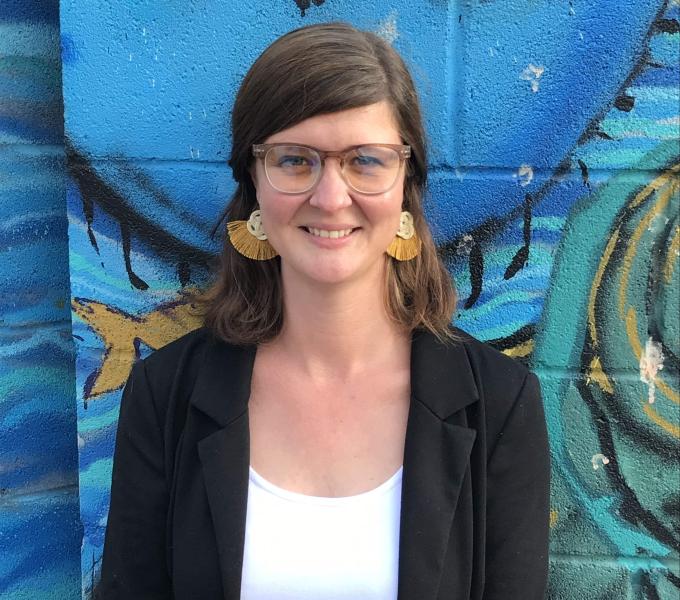 White woman with brown hair a white top, black blazer, yellow bottoms and yellow earrings standing in front of a blue mural background