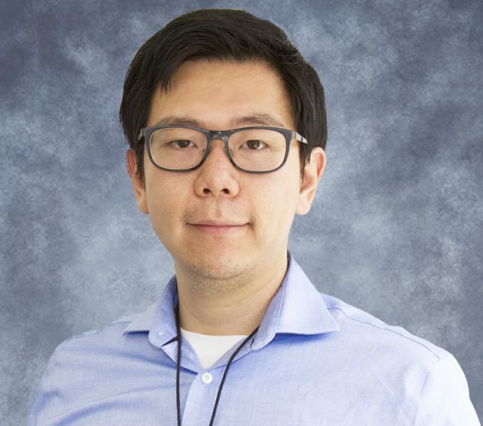 Asian male with black hair wearing a blue button up with a necklace wearing glasses.